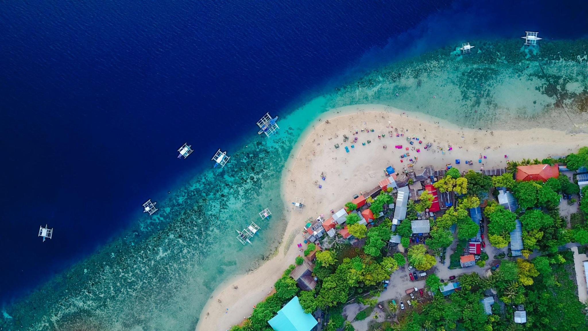 Luchtfoto van een tropisch strand met turquoise water, kleine bootjes langs de kust en kleurrijke parasols, omringd door weelderige groene bomen en huisjes (foto: Tirachard Kumtanom)