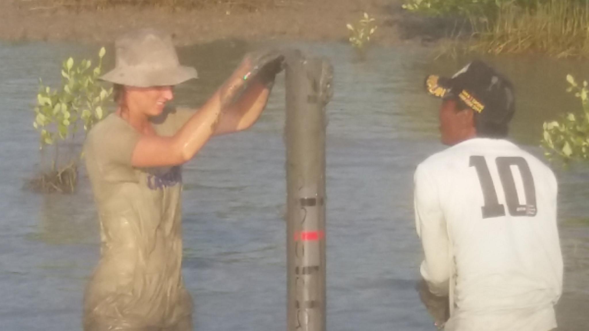 Twee mensen staan tot hun knieën in modderig water en zetten een verticale meetbuis of paal neer; de ene draagt modderige werkkleding en een hoed, en de andere draagt een wit shirt met het nummer 10 op de rug.