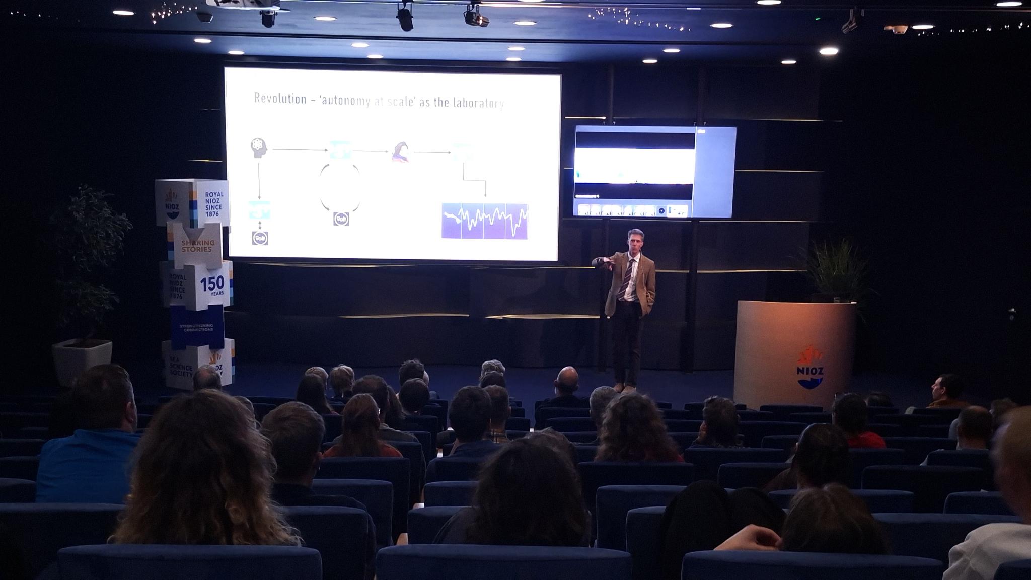 Een spreker die op het podium een lezing houdt voor een volle zaal tijdens een NIOZ-colloquium.