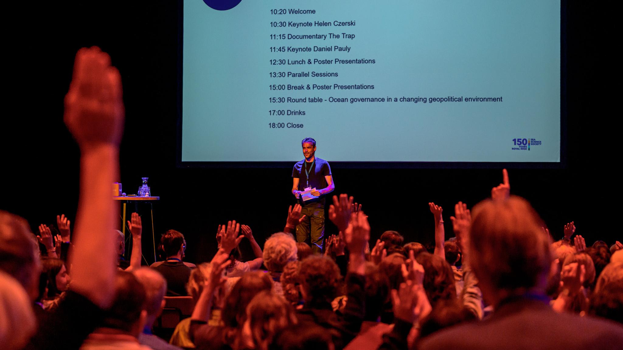 moDe presentator heet het publiek welkom op een podium voor een groot scherm derator welcomes audience on a stage in front of large screen