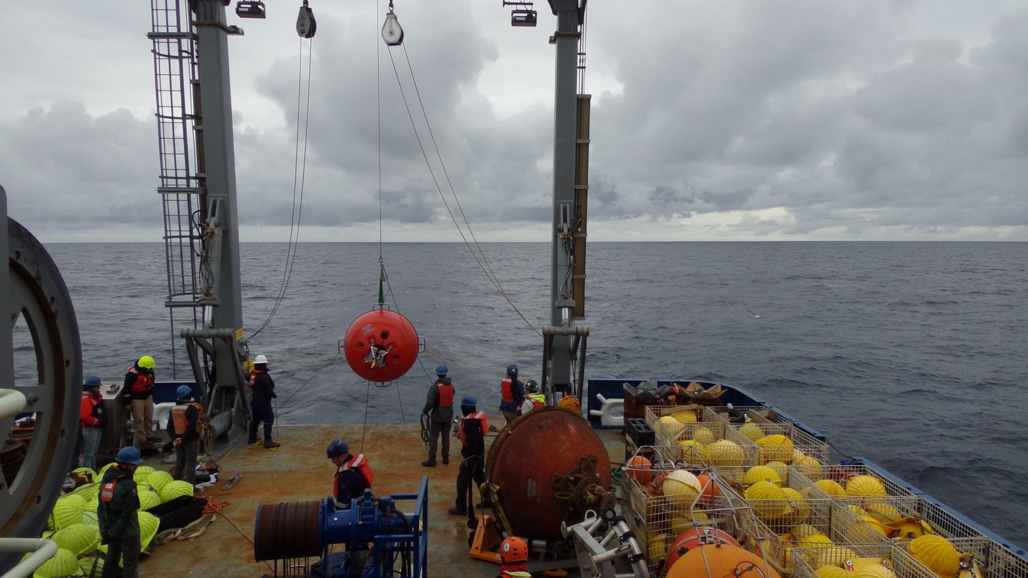 The rear deck of a vessel, where the A-frame crane hoists a large red ball that is the mooring. Crewmembers are standing by to reel it in.