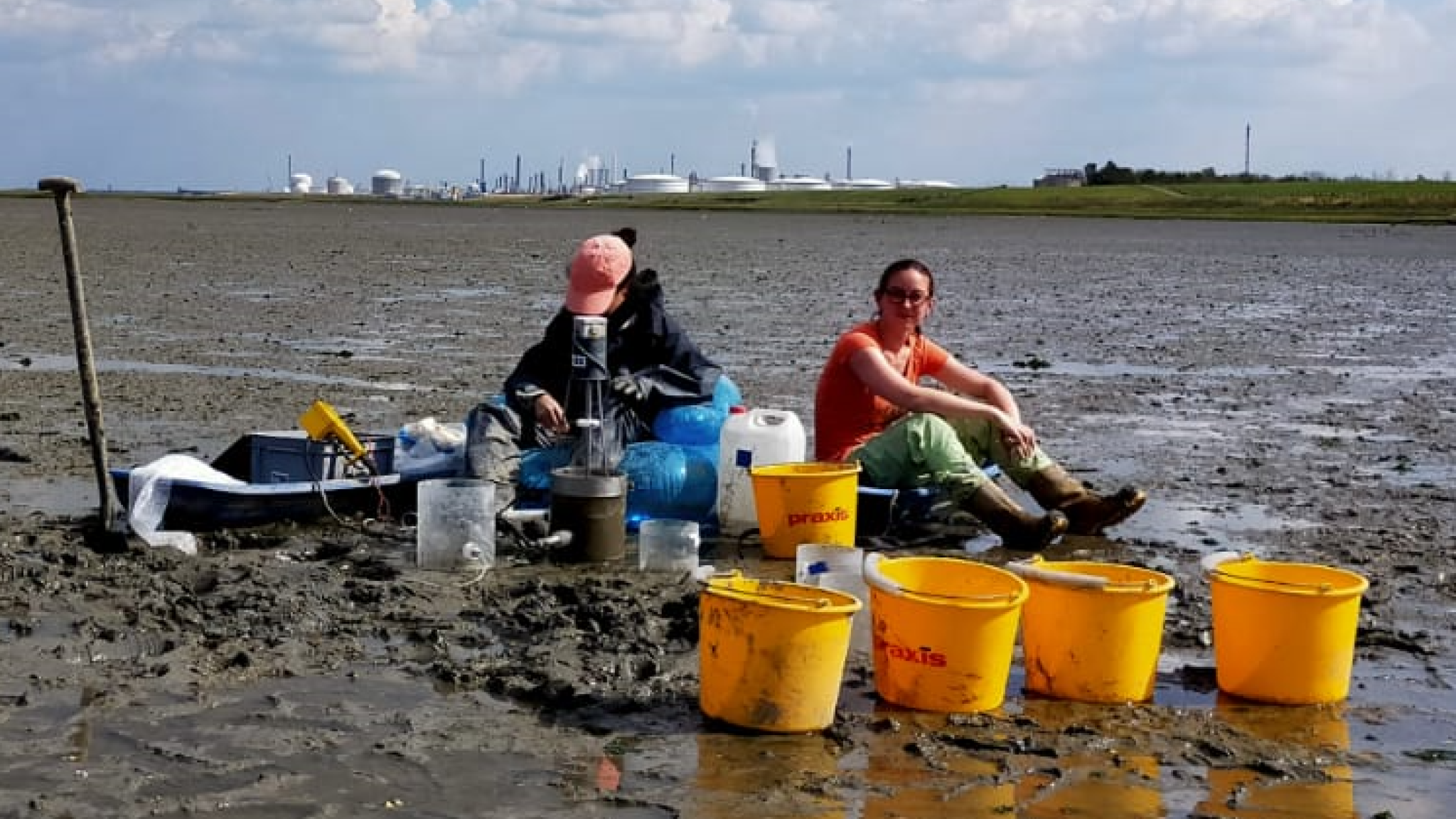 Researchers Alena di Primio and Dunia Rios-Yunes collect water samples during an erosion experiment on the tidal flats of the Oosterschelde. Sampling equipment and yellow buckets are arranged on the exposed mudflat, with an industrial area visible in the distance.