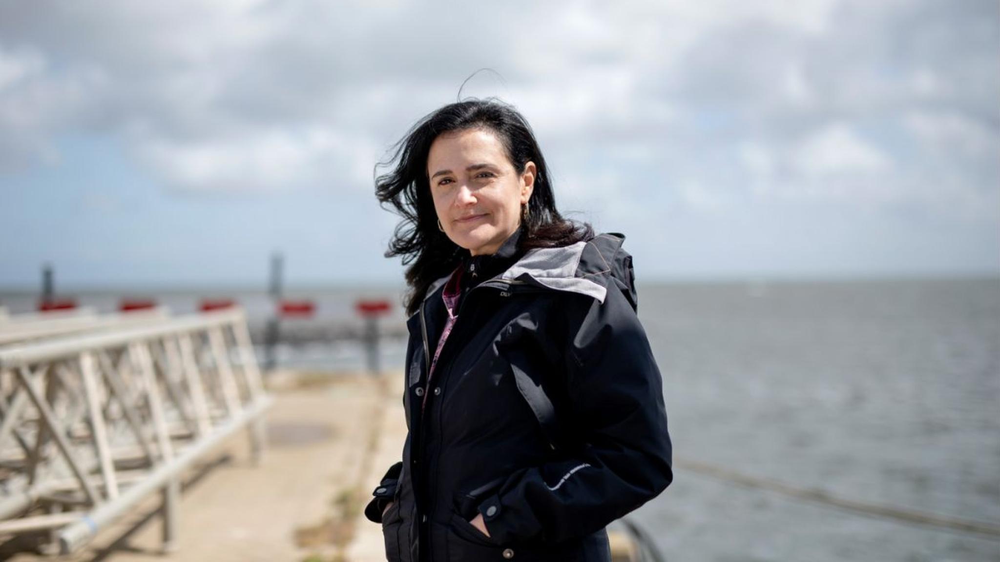 Portrait of female scientist in a dark coat standing on a pier with the sea and a cloudy sky in the background.