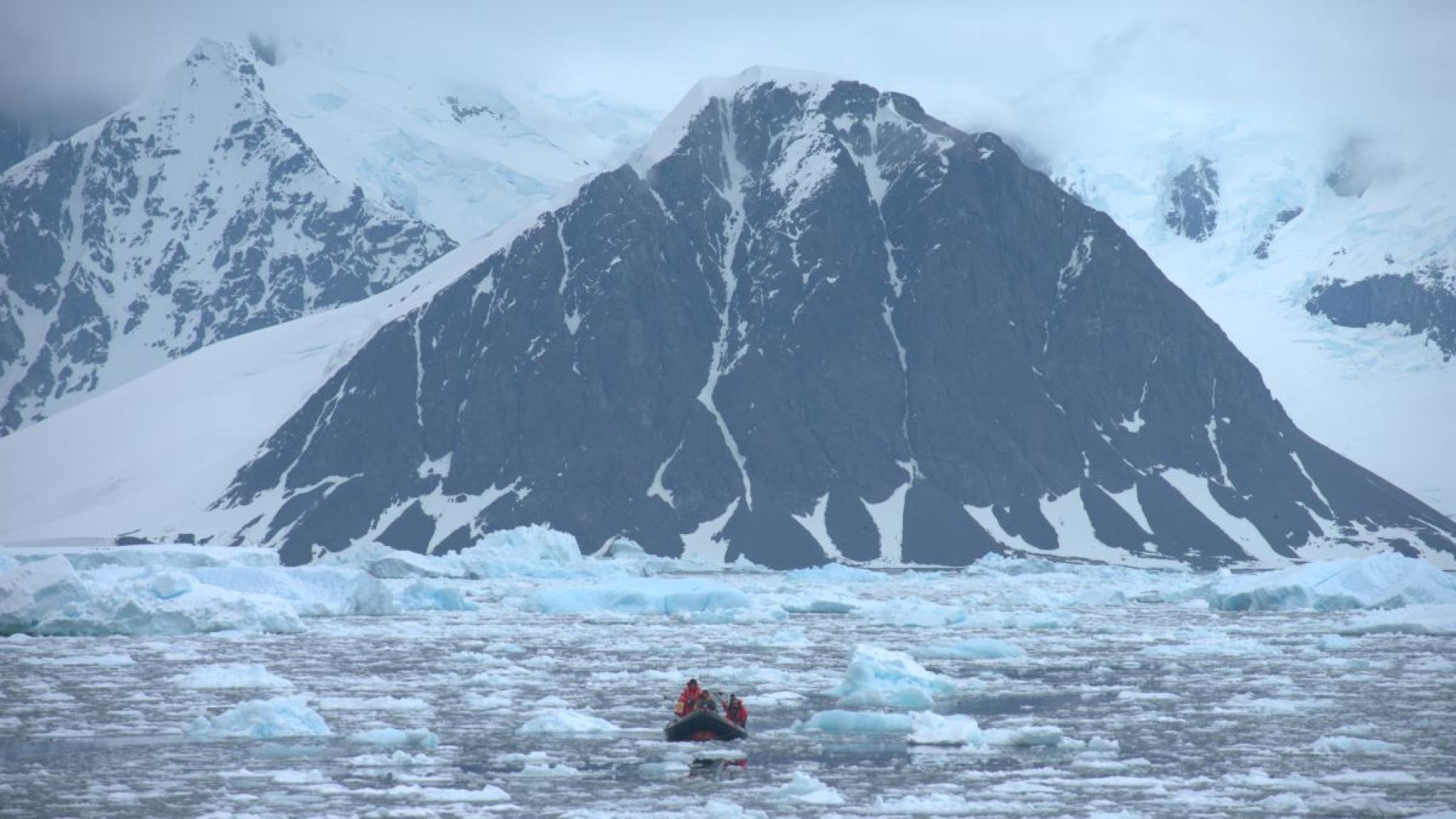 A small inflatable boat carrying researchers moves through icy water scattered with floes, heading toward a towering, snow-covered mountain with dark rock faces under a cloudy Antarctic sky.