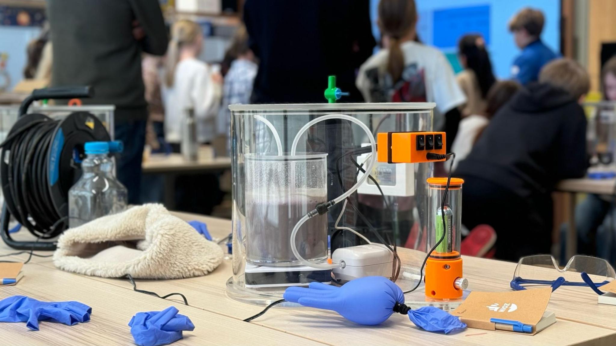 A demonstration setup with a clear tank and sensors on a table, surrounded by lab gloves and equipment, while people watch in the background.