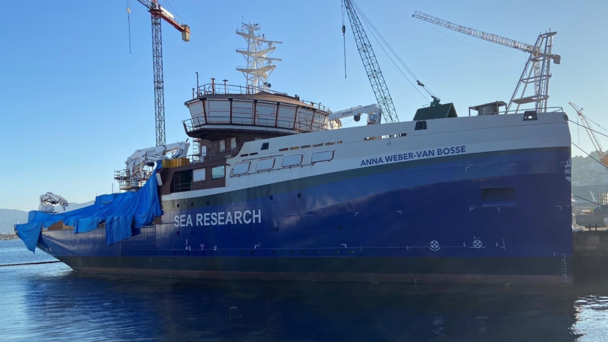 The ship RV Anna Weber van Bosse being under construction, seen from the side. The back deck is covered with palstic, and there are 3 cranes behind the ship lifting supplies on deck.