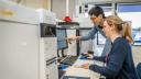 Two researchers looking at a computer, one pointing at the screen. The computer is connected to several laboratory machines, looking like big grey boxes atop a working bench.