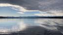Calm view across Lake Greifensee with gentle ripples on the water, dramatic cloudy skies, and snow-capped mountains on the horizon.