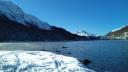 Snow-covered shoreline of Lake St. Moritz with clear blue water, surrounded by snow-capped mountains beneath a bright blue sky.