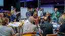 Een groep zittende mensen in een grote zaal luistert naar een staande man in een groen jasje