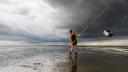Emma Penning standing on the mudflats wearing diving shoes and shorts, holding a long pole in the air with a metal sieve attached to it