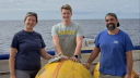 Three people standing on deck of a research vessel holding a large yellow buoy