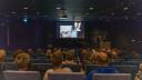 Lezing gezien vanuit het publiek in een donker verlichte zaal