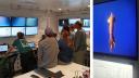 On the left: group of people watching live footage of under water images, to the right a large squid on screen