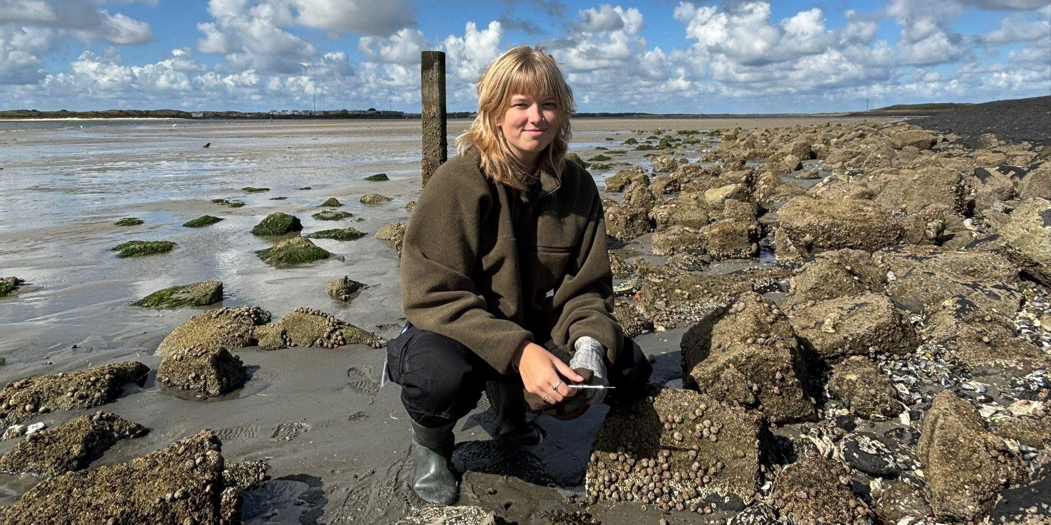 Rosalinde van Ooijen gehurkt op een rotsachtige kust tijdens laagwater, omringd door met zeepokken bedekte rotsen en plukken groen zeewier. Ze draagt een groene jas, zwarte broek en rubberlaarzen, met een deels bewolkte lucht en drooggevallen wadden die zich in de verte uitstrekken.
