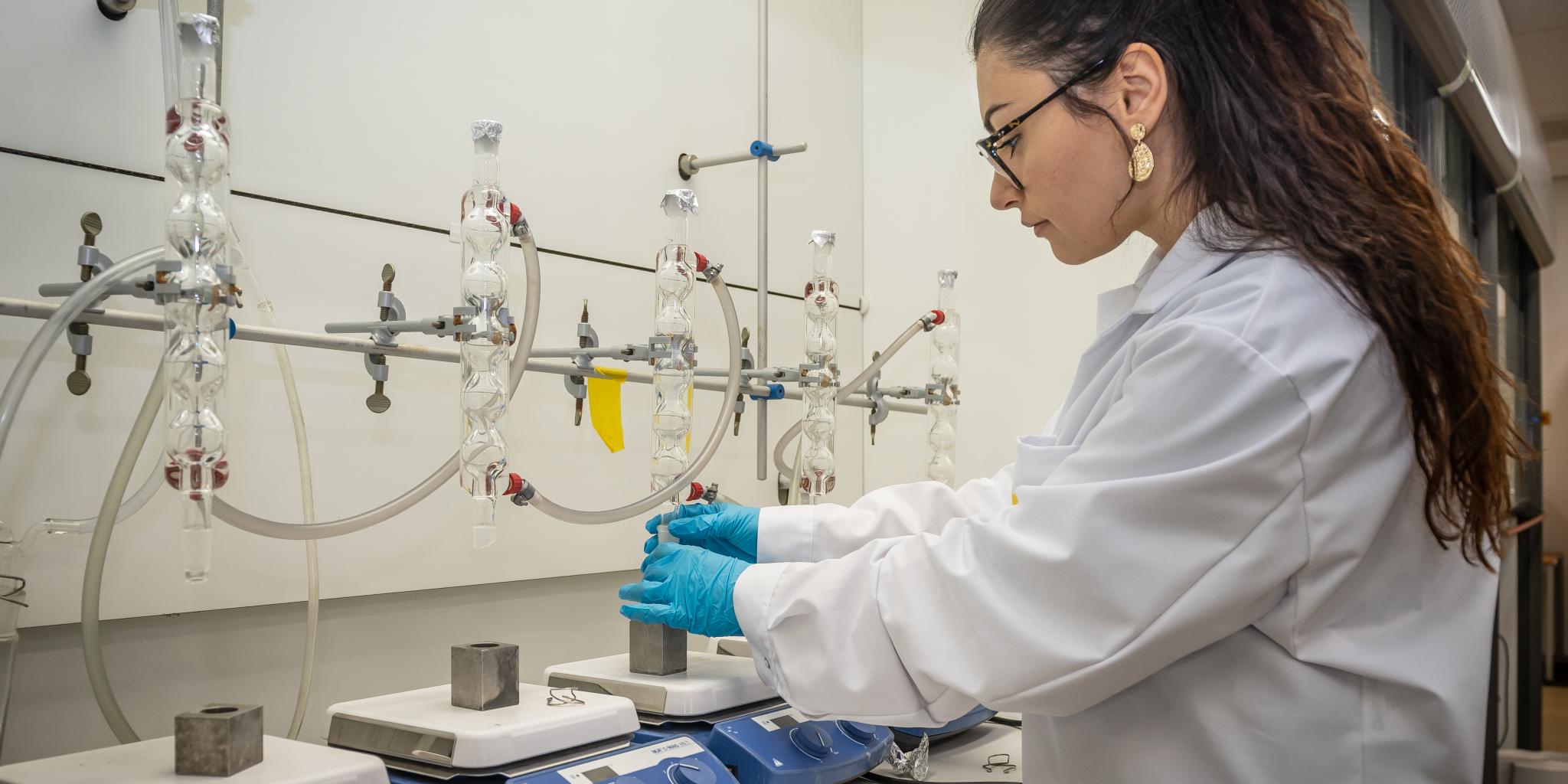 Een vrouw werkend in een laboratorium. Voor haar een werkbank met vier weegschaalachtige machines, elk met een uitgehold ijzeren blok waarop een monster geplaatst kan worden. Boven elk blok een glazen buisstructuur, waaruit monsters komen.