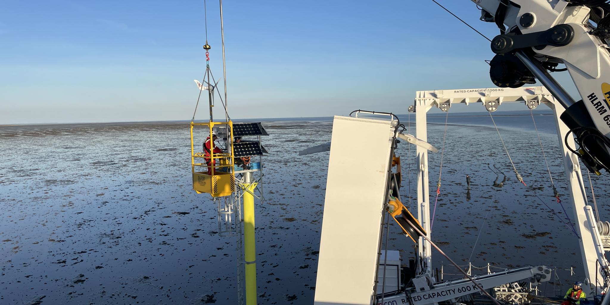 A crane, based on the deck of a ship, is holding a lift with a person in it next to an iron pole that has been placed in the sea bottom. The person is installing a solar panel atop the pole