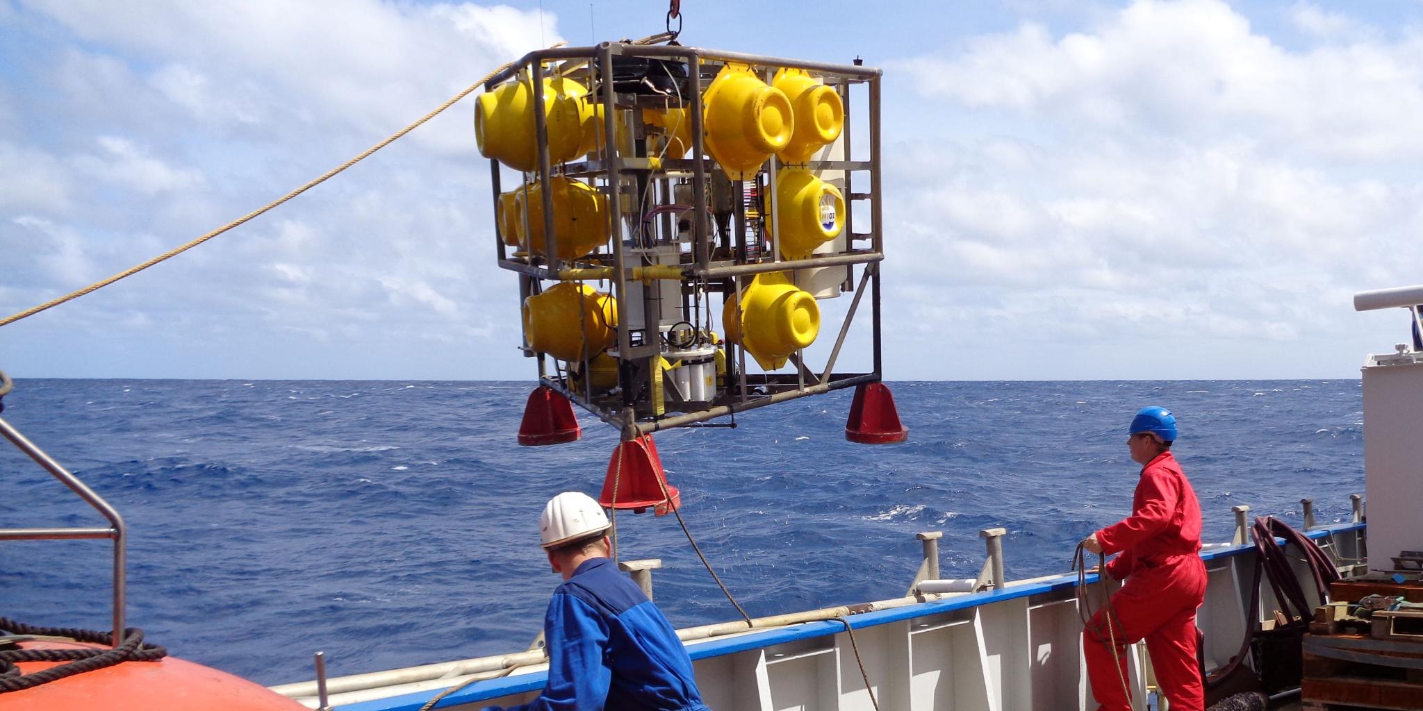 Two people on the deck of a research vessel. An iron frame with large yellow balls in it is lifted from the deck to be lowered into the ocean