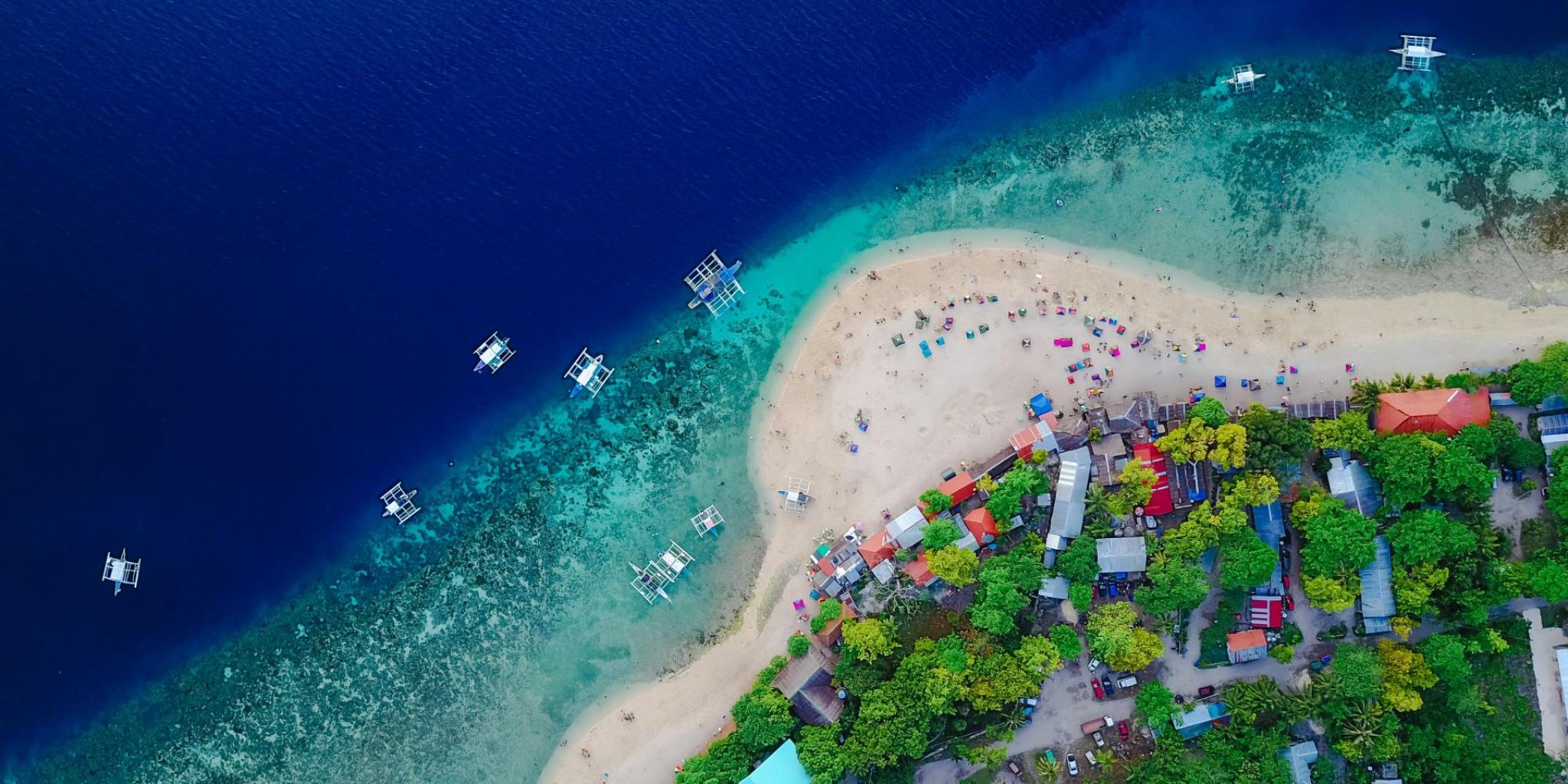 Aerial photo of a tropical beach with turquoise water, small boats along the coast and colourful parasols, surrounded by lush green trees and small houses (photo: Tirachard Kumtanom)