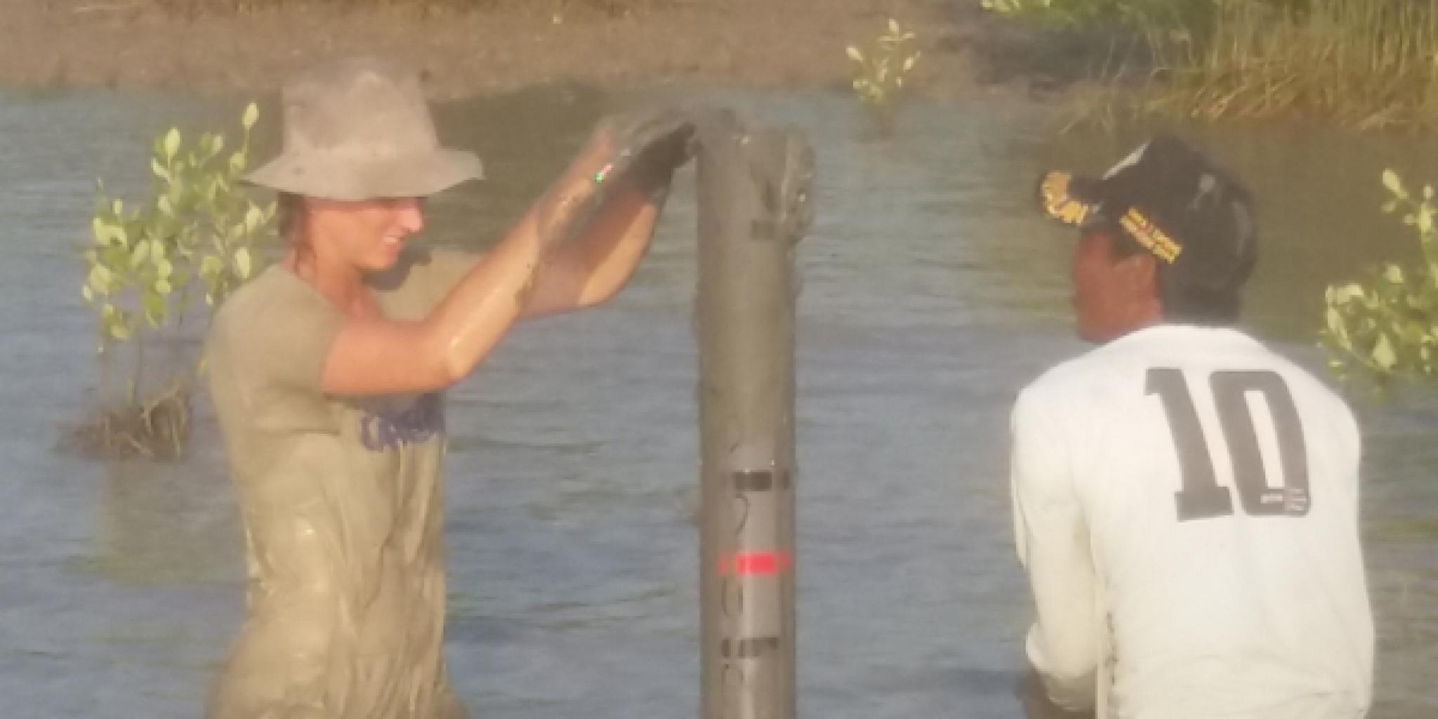 Twee mensen staan tot hun knieën in modderig water en zetten een verticale meetbuis of paal neer; de ene draagt modderige werkkleding en een hoed, en de andere draagt een wit shirt met het nummer 10 op de rug.