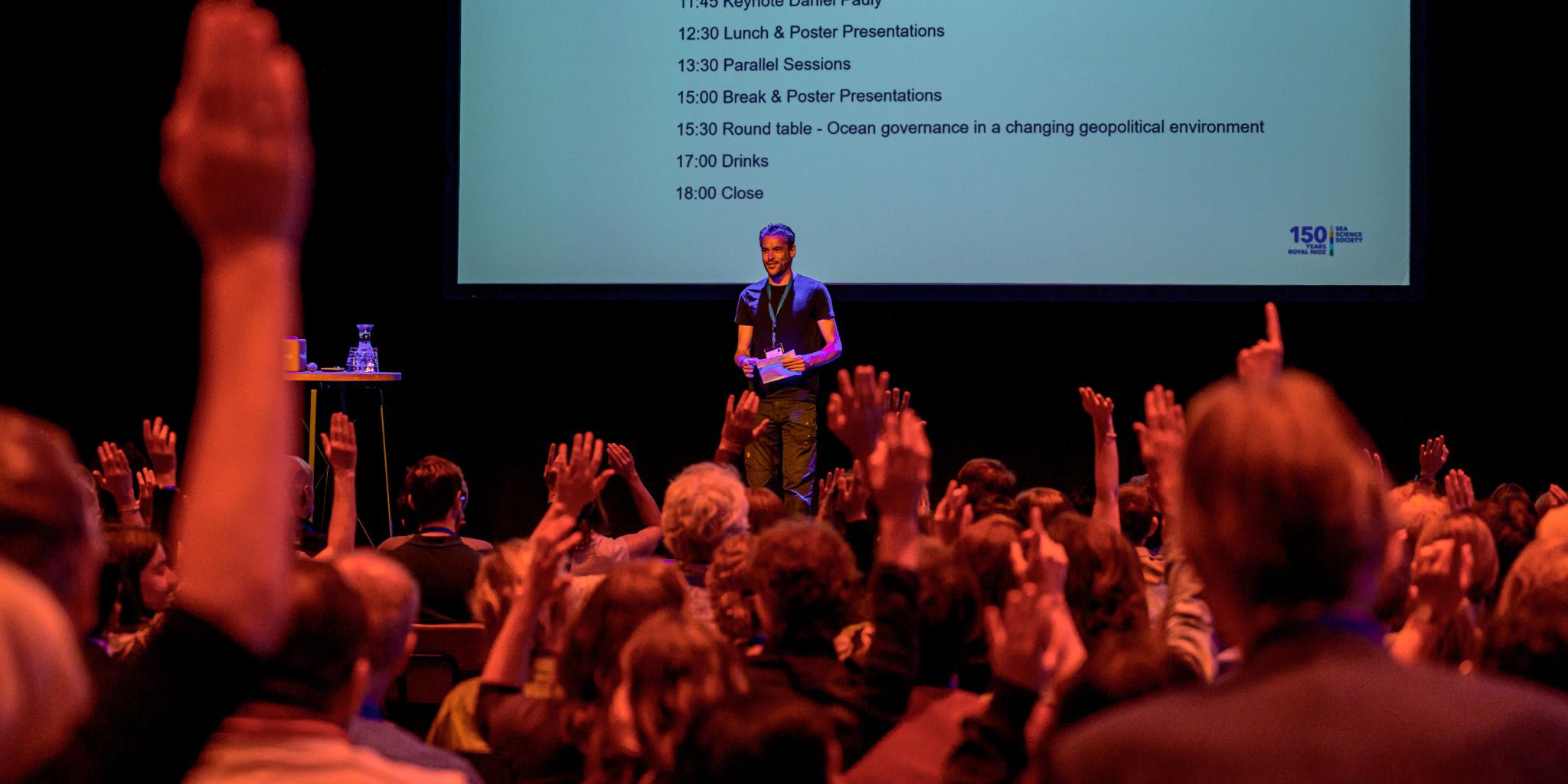 moDe presentator heet het publiek welkom op een podium voor een groot scherm derator welcomes audience on a stage in front of large screen