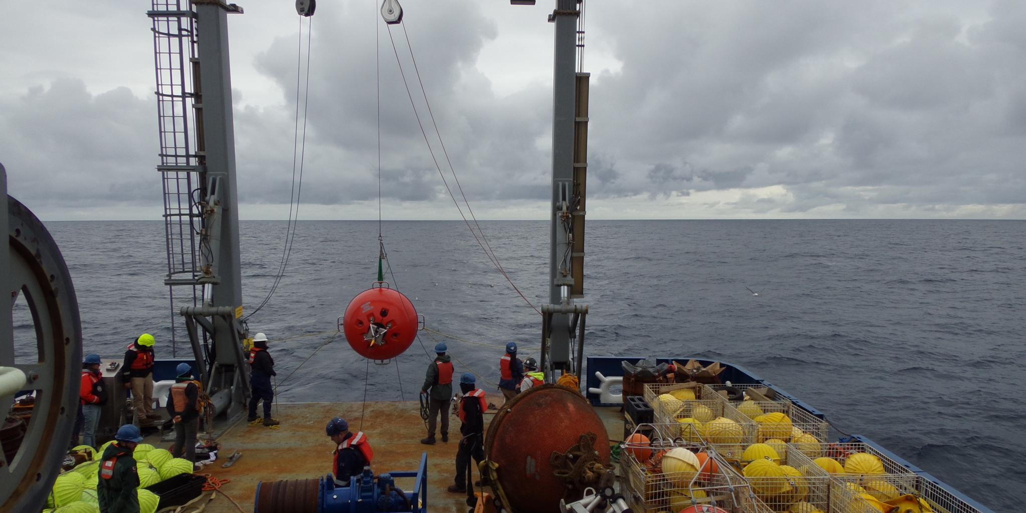 Het achterdek van een schip, waar de A-frame-kraan een grote rode bal omhoog hijst die als meerlijn dient. Bemanningsleden staan klaar om deze binnen te halen.