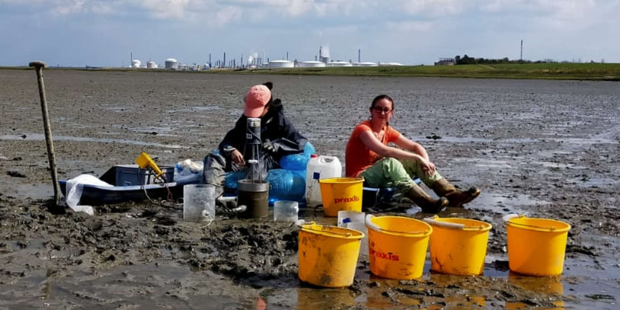 Researchers Alena di Primio and Dunia Rios-Yunes collect water samples during an erosion experiment on the tidal flats of the Oosterschelde. Sampling equipment and yellow buckets are arranged on the exposed mudflat, with an industrial area visible in the distance.