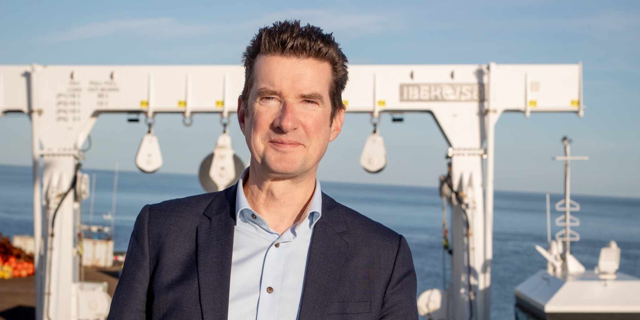 Glimlachende man op het dek van een onderzoeksschip met een groot wit frame en de zee op de achtergrond