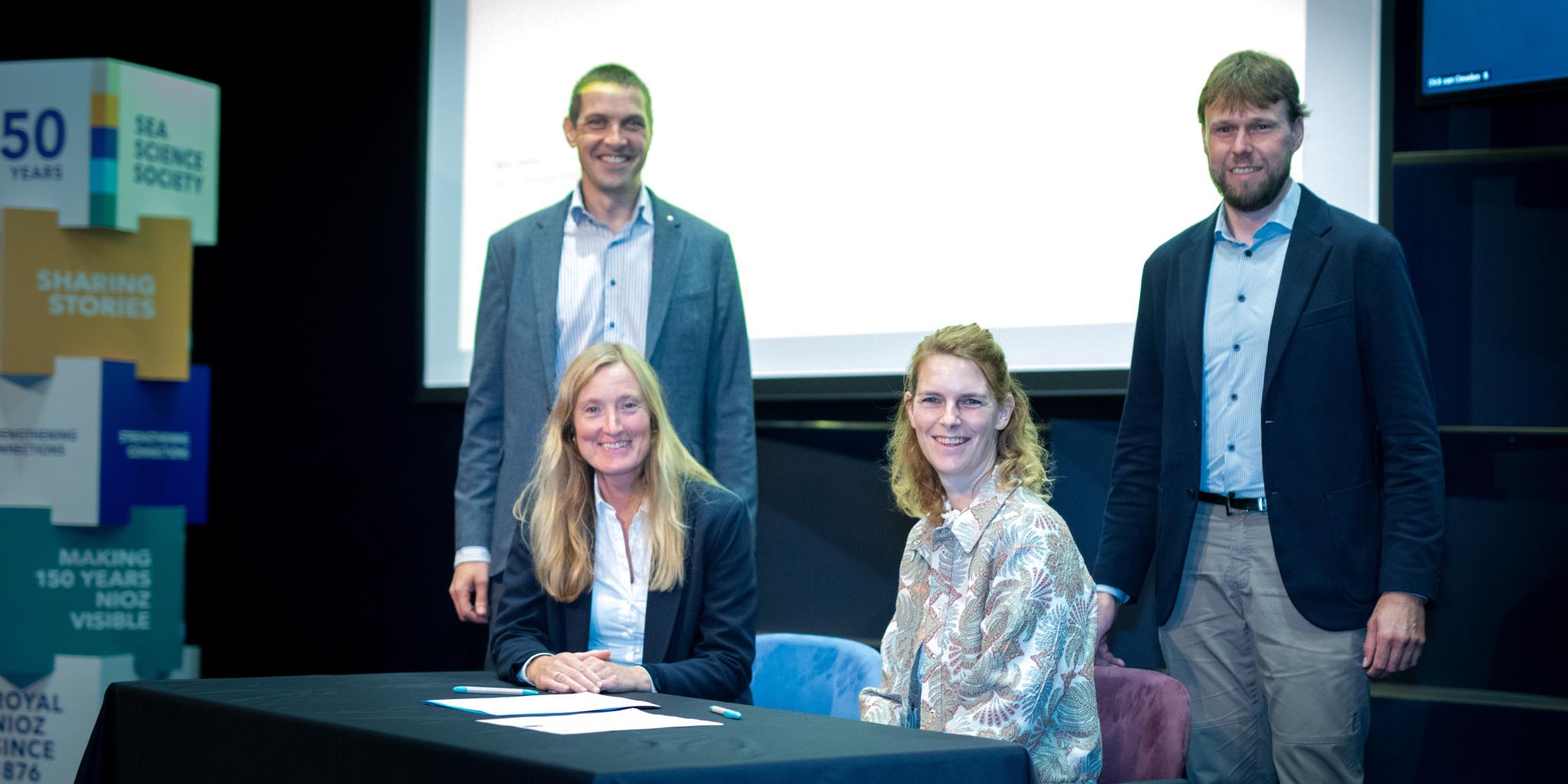 Twee glimlachende vrouwen die achter een tafel zitten, hebben zojuist papieren ondertekend. Achter hen staan twee mannen voor een wit scherm.