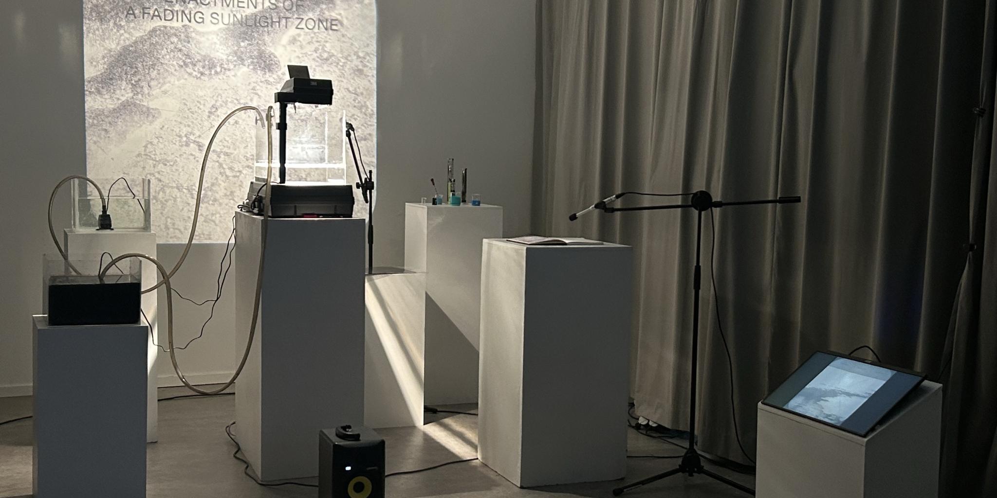 A museum like set up with several white columns and a screen in the background. On one pillar is a large transparent box filled with water that is lit from above, another holds a screen and a third the box of darker fluid that will be pumped into the clear water.