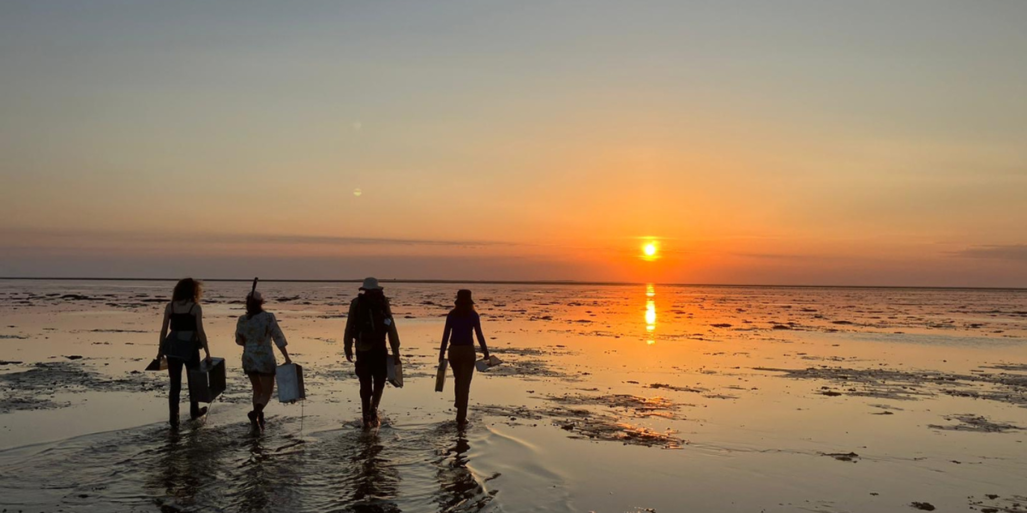 Vier mensen lopen over een ondiep strand bij zonsondergang; hun silhouetten worden weerspiegeld op het natte zand.