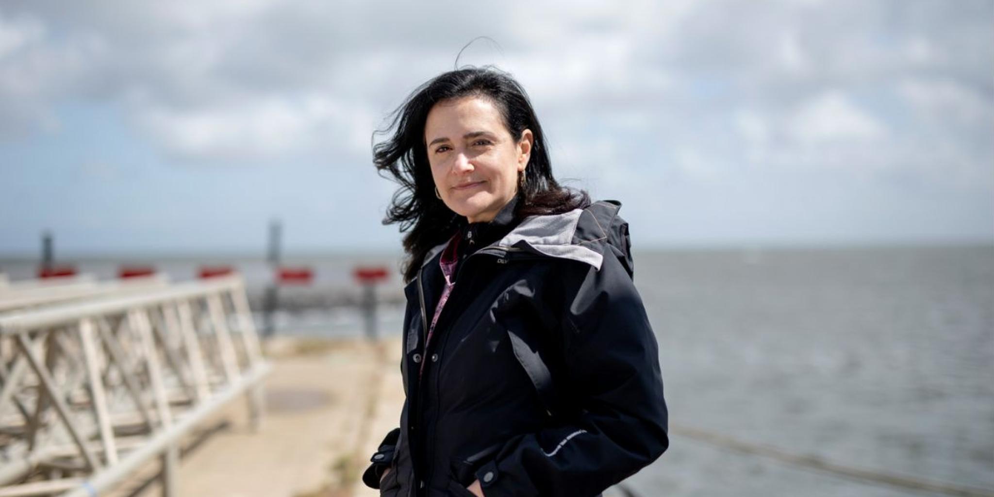 Portrait of female scientist in a dark coat standing on a pier with the sea and a cloudy sky in the background.