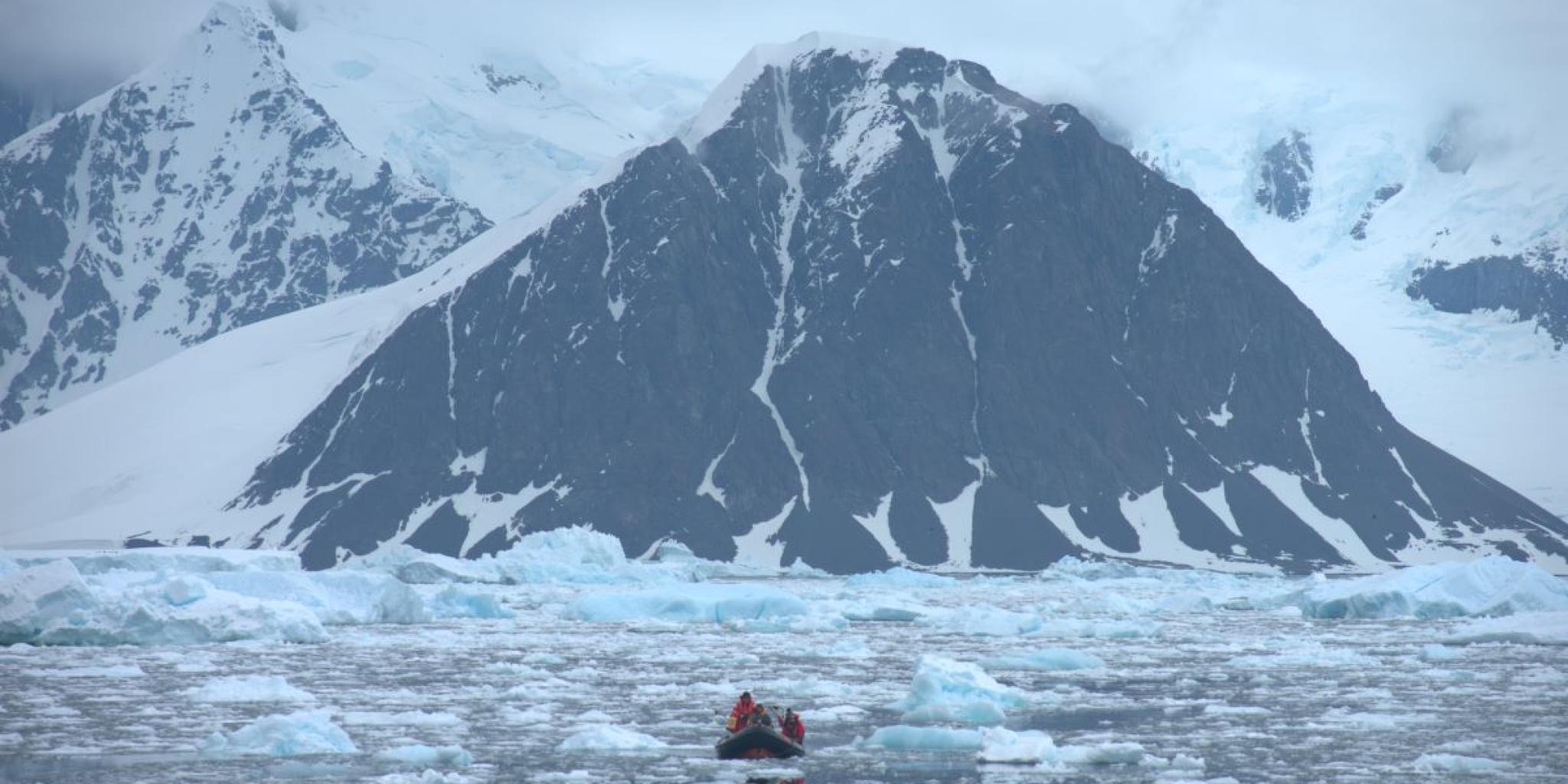 A small inflatable boat carrying researchers moves through icy water scattered with floes, heading toward a towering, snow-covered mountain with dark rock faces under a cloudy Antarctic sky.