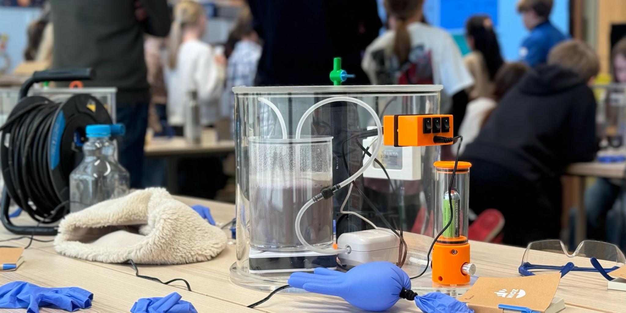 Een demonstratieopstelling met een heldere bak en sensoren op een tafel, omringd door labhandschoenen en apparatuur, terwijl mensen op de achtergrond toekijken.