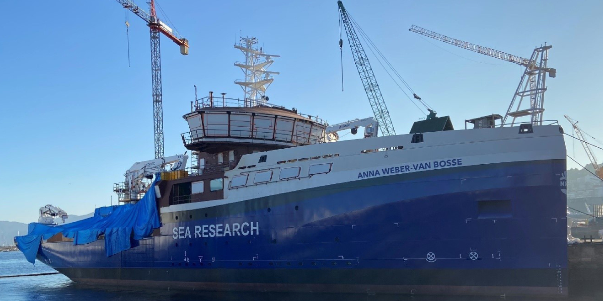 The ship RV Anna Weber van Bosse being under construction, seen from the side. The back deck is covered with palstic, and there are 3 cranes behind the ship lifting supplies on deck.