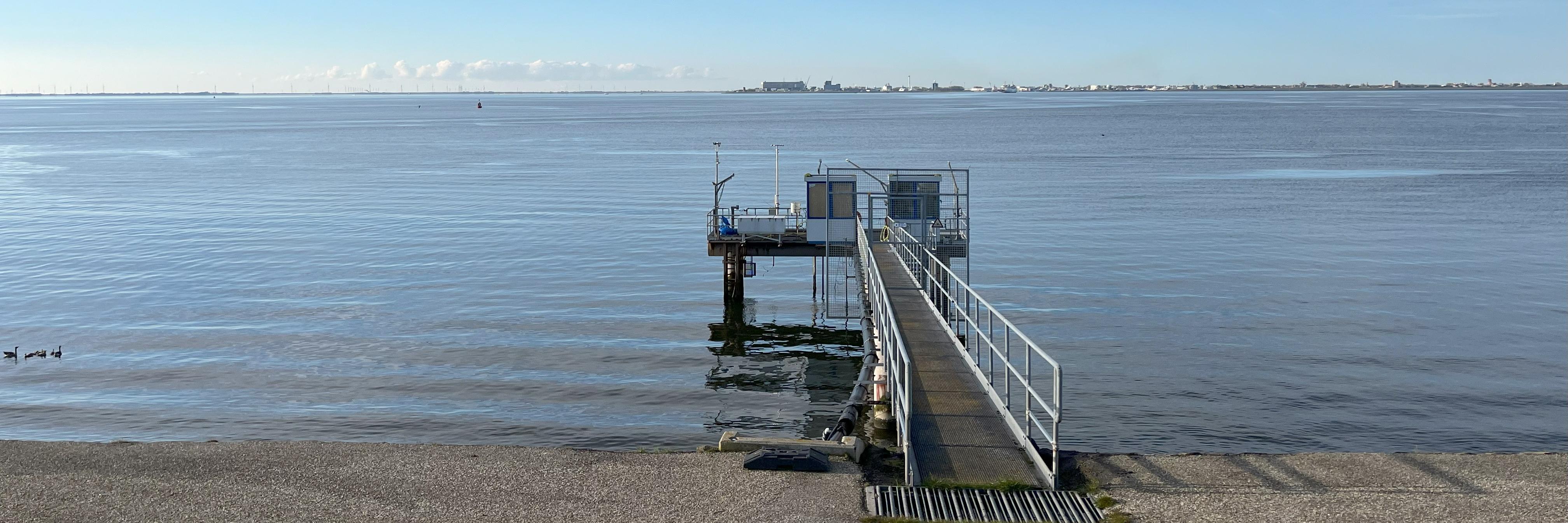A small metal jetty protrudes from a dyke into the water. On the jetty is a small cabin and several measurement set ups can be seen.