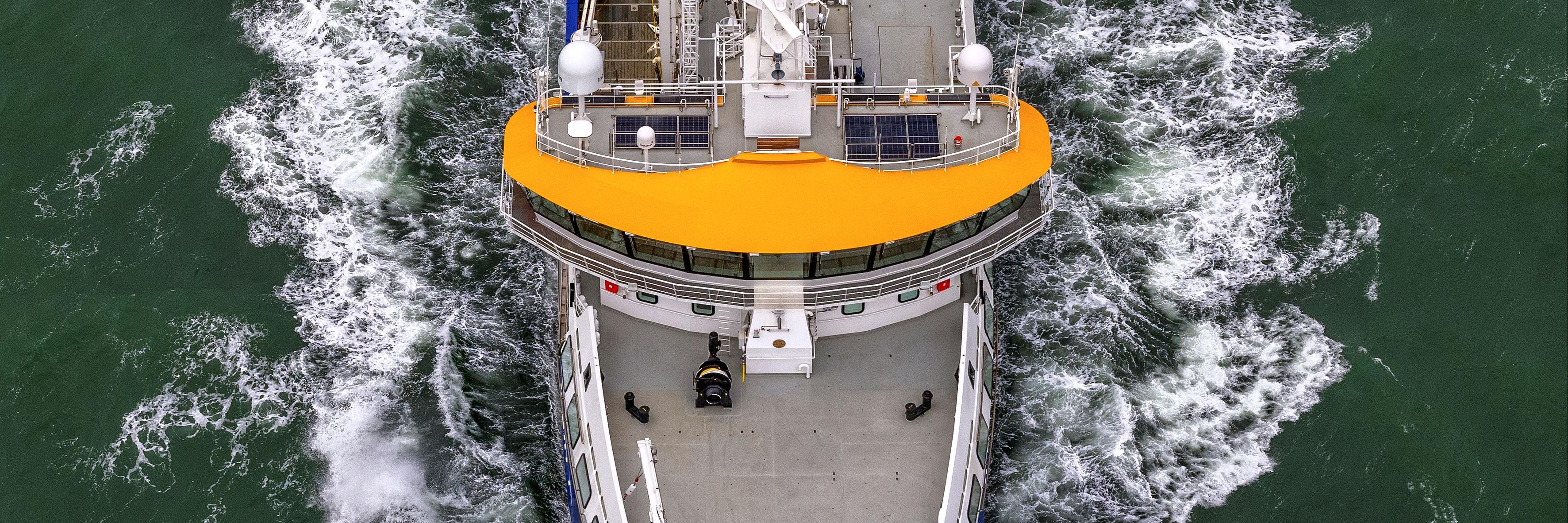 An aerial view of a research vessel moving through the water, with scientific equipment and crew visible on deck.