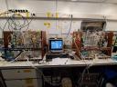 Op een werkbank staat een laboratoriumopstelling met twee complexe glazen bioreactorsystemen die via slangen, sensoren en bedrading met elkaar zijn verbonden, naast een computer die wordt gebruikt voor het verzamelen en monitoren van gegevens. De opstelling is ontworpen voor gecontroleerde experimenten onder laboratoriumomstandigheden.