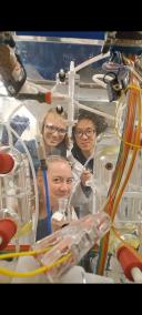 Drie onderzoekers glimlachen voor een opstelling van glazen laboratoriumreactoren, slangen en wetenschappelijke apparatuur in een marien onderzoekslaboratorium. De complexe proefopstelling vult de voorgrond, terwijl de onderzoekers op de achtergrond te zien zijn.