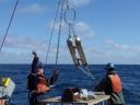 Crew members aboard the research vessel use a hook to catch the yellow buoy of a recovered ocean mooring while waves roll alongside the ship.