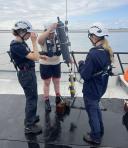 Three students wearing life jackets and helmets prepare a Niskin bottle to collect surface water samples on the deck of the research vessel HA1-Wantij.