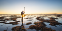 A researcher stands on an intertidal mussel cultivation plot in the Oosterschelde at low tide, holding a GPS survey pole. The setting sun shines through the figure, illuminating the exposed mussel beds and shallow pools.