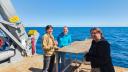 Expedition participants enjoying sunny weather together on the deck of the research vessel with the open ocean in the background.