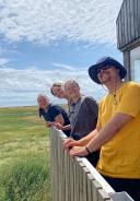 Vier leden van het onderzoeksteam staan op het balkon van hun huisje en kijken uit over het groene landschap en de zee.