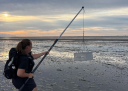 Femke Vermeulen loopt over het wad en draagt met een lange stok een metalen bemonsteringsapparaat voor garnalen.