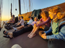 Een groep mensen zit ontspannen bij elkaar op het dek van zeilschip De Ideaal in het warme licht van de ondergaande zon.