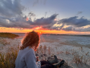 Een persoon zit op het strand en kijkt naar een feloranje zonsondergang boven de Waddenzee, met donkere wolken aan de hemel.