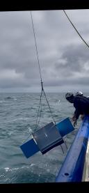 Crew members wearing safety helmets guide a manta net suspended by a crane above the rough sea.