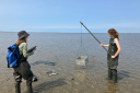 Two people in waders stand in shallow water, one holding a clipboard and the other lowering a metal sampling box on a pole into the sea.
