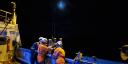 Researchers in helmets and orange safety gear handle sampling equipment on the deck of a ship at night under the moonlight.